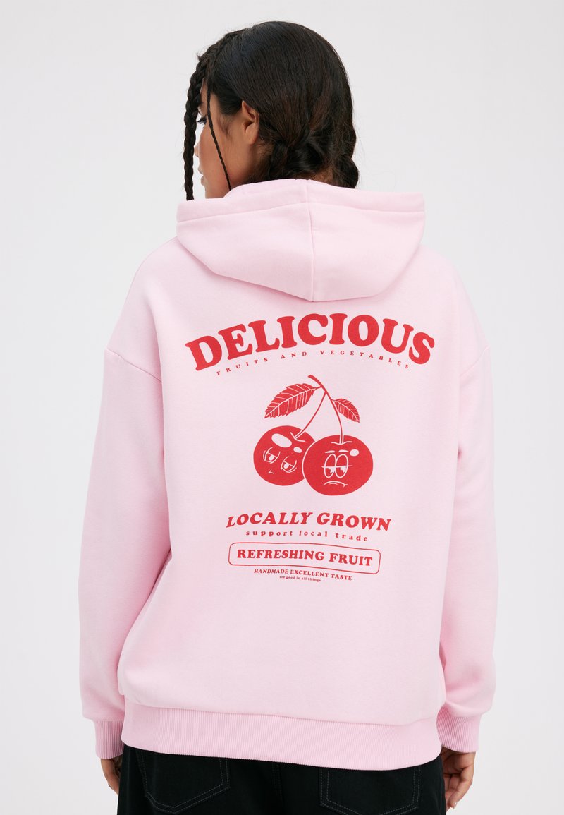 Sweat à capuche rose avec un graphique rouge représentant des cerises, le texte indique « DÉLICIEUX », « CULTIVÉ LOCALMENT » et « FRUIT RAFFRAÎCHISSANT ». Matériau en coton doux.