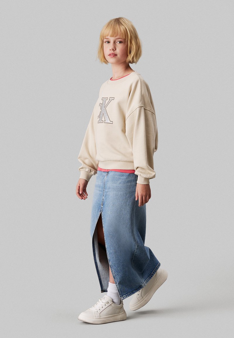 Sudadera de gran tamaño en color crema con el logo "SK", combinada con una falda de mezclilla azul clara con una abertura en la parte delantera y zapatillas blancas.
