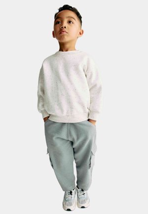 Felpa grigia chiara con motivi a macchie e vestibilità morbida, abbinata a pantaloni cargo grigi chiari con texture. Sneakers sportive bianche e blu completano il look.