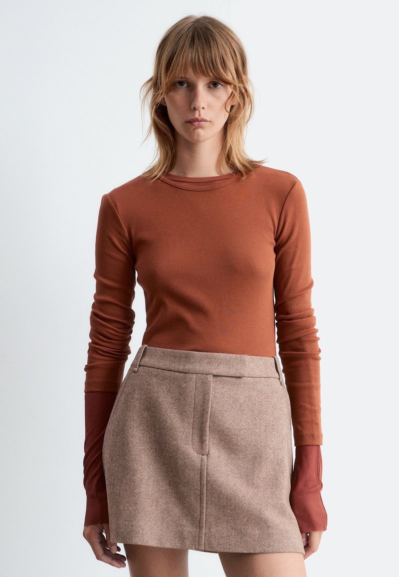 Langärmliges geripptes Oberteil in Rostfarbe mit Rundhalsausschnitt, kombiniert mit einem hellbraunen Rock mit strukturiertem Finish und frontalen Taschen.