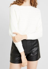 Pull tricoté blanc à côtes avec un large décolleté et des manches longues, associé à un short en cuir noir orné de détails de couture décorative.