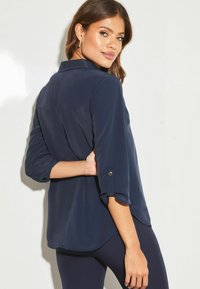 Blusa leggera di colore blu navy, con colletto, maniche tre quarti arrotolate sui polsini e orlo curvo.