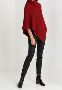 Femme portant un poncho rouge en tricot avec col châle, un pantalon skinny noir et des bottines noires brillantes, debout devant un fond uni.