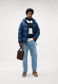 Jachetă puffer bleumarin cu glugă cu fermoar, combinată cu un pulover alb și bleumarin cu dungi, blugi deschiși și o geantă maro. Pantofii din suede completează ținuta.
