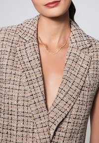 Beżowa kamizelka w kratę z głębokim dekoltem w kształcie litery V, teksturowana tkanina z tkanymi wzorami w brązowym i czarnym kolorze. Akcent w postaci złotego łańcuszka na szyi.