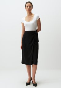 Zwarte wikkelrok van textuurstof, met een strik in de taille en een kleine uitsparing. Gecombineerd met een aansluitend wit T-shirt met korte mouwen en hoge hakken.