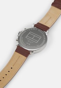 Montre Tommy Hilfiger avec un boîtier en acier inoxydable, fond gris, bracelet en cuir véritable marron et détails de logo gravé. Résistante à l'eau jusqu'à 5 bars.