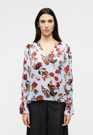 Γυναίκα με μακριά σκούρα μαλλιά, ντυμένη με ανοιχτό γαλάζιο μπλουζάκι με κόκκινο λουλουδάτο σχέδιο και μαύρο παντελόνι, στέκεται μπροστά σε ένα απλό φόντο.