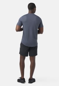 Uomo in piedi di spalle che indossa una maglietta grigia a maniche corte con trama, pantaloncini sportivi neri e scarpe da ginnastica nere su sfondo bianco.