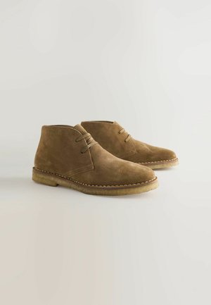 Brown suede ankle boots with rounded toes, two eyelets for laces, and stitched detailing along the tan rubber sole.