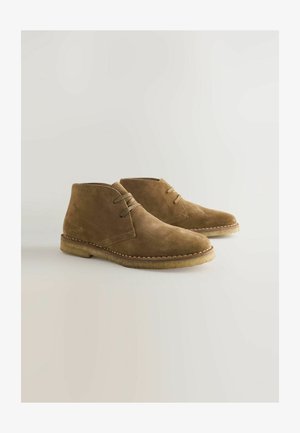 Bocanci scurți din suede maro, cu vârfuri rotunjite, două găuri pentru șireturi și detalii cusute pe talpa de cauciuc bej.