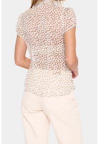 Camicia leggera e trasparente con collo alto arricciato, maniche corte e motivo a pois in tonalità beige. Indossata con pantaloni chiari.