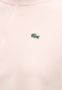 Gros plan d’un tissu texturé rose clair avec une fermeture éclair à l’avant et un petit logo crocodile vert brodé.