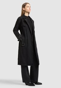 Fiatal nő áll hosszú, fekete, kétsoros gombos farmerkabátban, fekete bő szárú nadrágban, fekete csizmában és fekete felsőben, szürke háttér előtt.