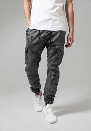 Cargo-Hosen im schwarzen Camouflage-Muster, mit elastischen Bündchen, Kordelzugbund und Seitentaschen. Kombiniert mit weißen Sportschuhen.