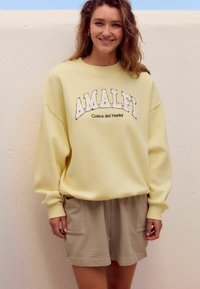 Gelbes Sweatshirt mit schwarzer "AMALFI" Stickerei, lockere Passform. Kombiniert mit beigen strukturierten Shorts mit seitlichen Taschen.