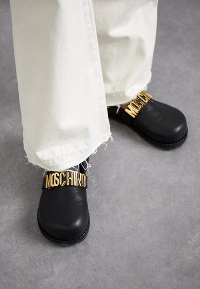 Μαύρες δερμάτινες παπούτσια κλεισίματος με χρυσά γράμματα "MOSCHINO". Στρογγυλή μύτη, χοντρή σόλα και απλός σχεδιασμός σε συνδυασμό με λευκά τζιν με φθορές.