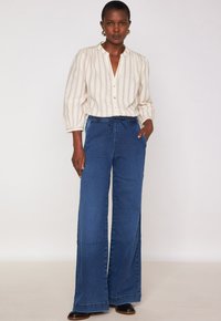 Donna in piedi che indossa una blusa a righe crema con chiusura a bottoni, jeans larghi blu con cintura a corda, scarpe nere e orecchini a cerchio dorati.