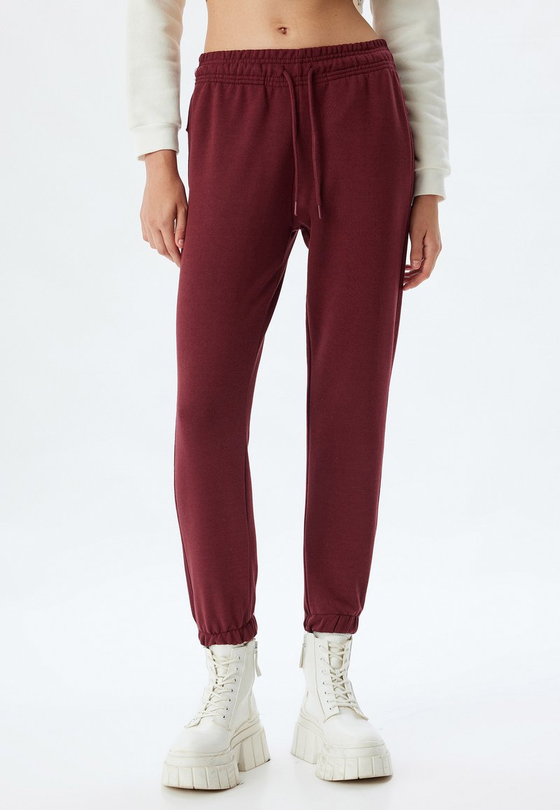 Mørkerøde sweatpants har en elastisk talje med snor, manchetter ved anklerne og en blød tekstur, parret med klodsede hvide støvler.
