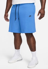 Man som bär ljusblå Nike-shorts med svarta snören, vita Nike-strumpor och vita Nike-sneakers, stående mot en vit bakgrund.