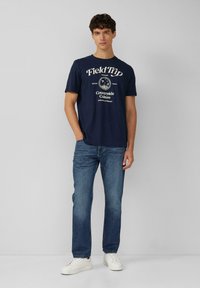 Navyblauwe katoenen T-shirt met witte bedrukte tekst en een grafisch ontwerp, gecombineerd met lichtblauwe denim jeans en witte sneakers.