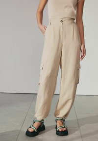 Beige cargo bukser med høj talje, med to sidelommer og knaplukning, parret med kraftige sorte sandaler og grønne accenter.