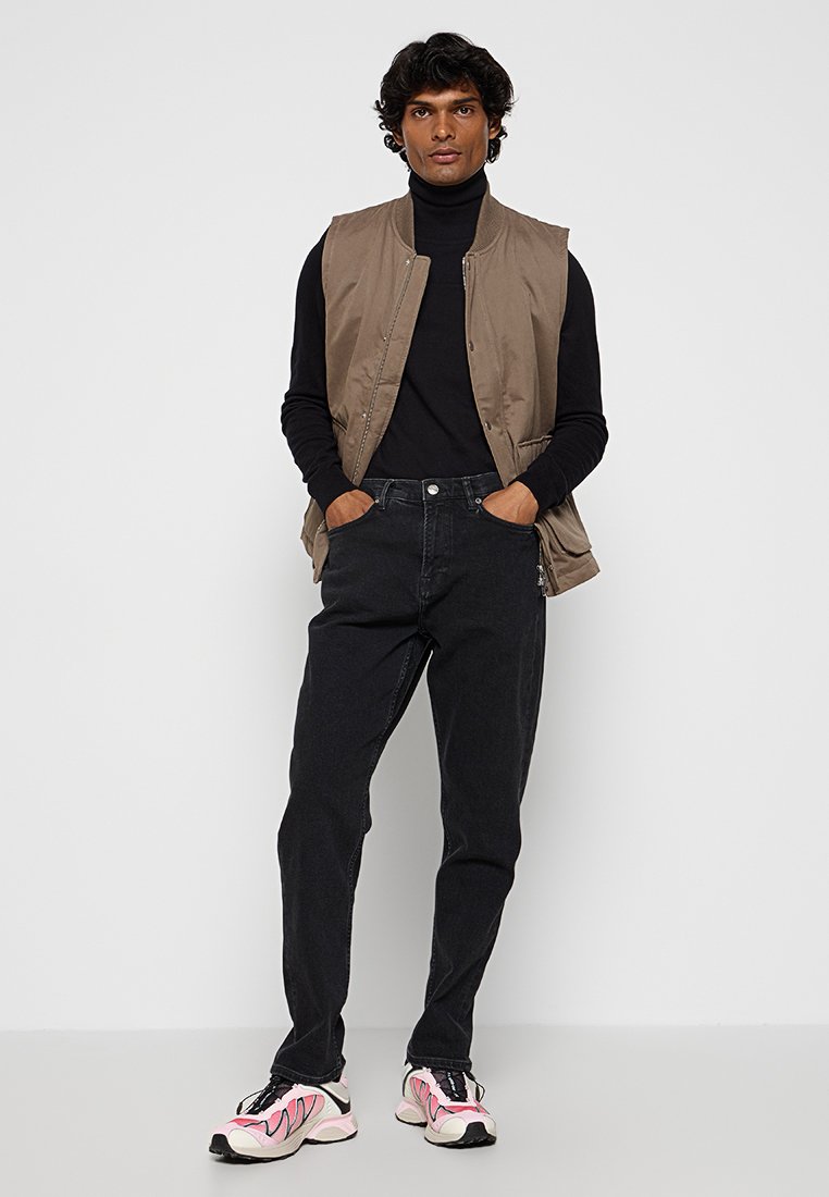 Khaki puchowa kamizelka na czarnym golfie, w zestawieniu z czarnymi dżinsami i trampkami z różowymi akcentami. Model stoi z rękami w kieszeniach.