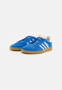 Modri športni čevlji iz semiša z belimi in rdečimi črtami, zaokroženi prsti, gumenimi podplati in ravnimi vezalkami, ki se ponašajo s klasičnim dizajnom.