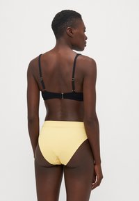 Svart ribbad bikinitopp med tunna band och guldfärgad detalj, kombinerad med högmidjade gula ribbade bikiniunderdelar. Bakvy.