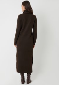 Brun turtleneck kjole lavet af blødt strikket materiale, med lange ærmer og sideslids, parret med knæhøje brune støvler for kontrast.