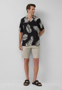 Zwarte kortärmelige shirt met wit bladerenpatroon, gecombineerd met beige shorts en zwarte sandalen. Minimalistisch ontwerp en relaxte pasvorm.