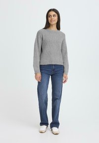 Szürke kötött pulóver, bársonyos nyakkivágással, sötétkék, széles nadrágú farmerrel kombinálva, fehér sportcipővel viselve. Sima textúra és bordás mintázat.