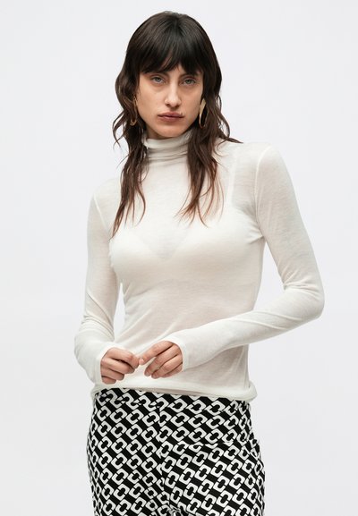 Λευκό μακρυμάνικο τοπ με ψηλό λαιμό από ελαφρύ ύφασμα, με εφαρμοστή σχεδίαση και θηλάκια για τον αντίχειρα στα μανίκια, συνδυασμένο με ασπρόμαυρο παντελόνι με σχέδιο.