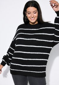 Frau mit langen dunklen Haaren, die einen schwarz-weißen gestreiften Strickpullover trägt, lächelt mit geschlossenen Augen und hebt einen Arm vor einem weißen Hintergrund.