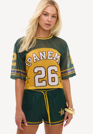 Femme blonde portant un maillot de sport vert et jaune avec le numéro 26 et un short assorti, debout avec une main baissée.