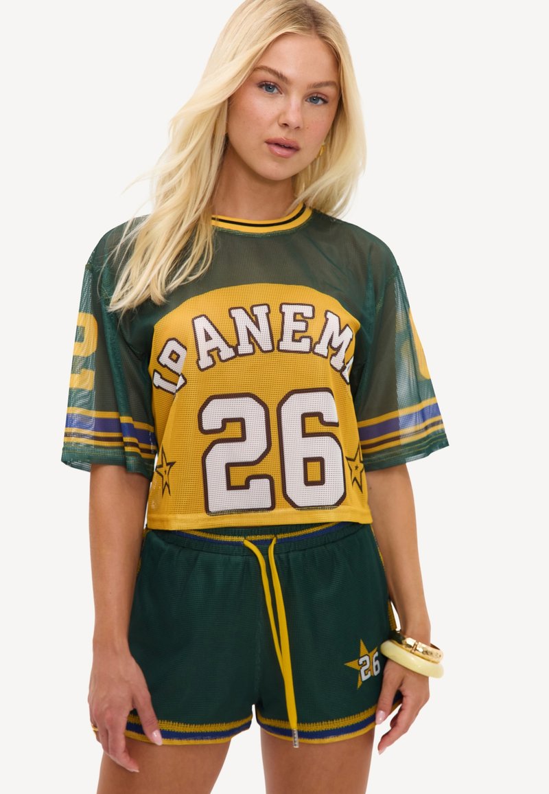 Femme blonde portant un maillot de sport vert et jaune avec le numéro 26 et un short assorti, debout avec une main baissée.