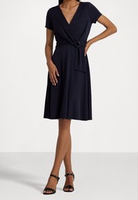 Abito a portafoglio blu navy con maniche corte, scollatura a V profonda, dettaglio con fiocco laterale e gonna lunghezza ginocchio. Tessuto morbido con leggera elasticità. Sandali neri con tacco.