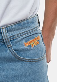 Los pantalones cortos de mezclilla de un azul claro presentan un logo bordado "homeboy x-tra" en naranja y amarillo en el bolsillo trasero. Diseño clásico de cinco bolsillos.