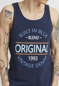 Tumesinine tanktop, mis on valmistatud puuvillast, esipaneelil trükk: "BUILT IN BLUE," "ORIGINAL" ja "VINTAGE DENIM," kaunistava tekstiga.