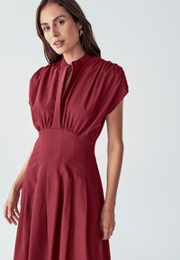 Červené šaty s vysokým golierom, krátkymi puff rukávmi a priliehavým tielkom. Tkanina je hladká s voľnou sukňou. Pozoruhodný detail gombíka na krku.