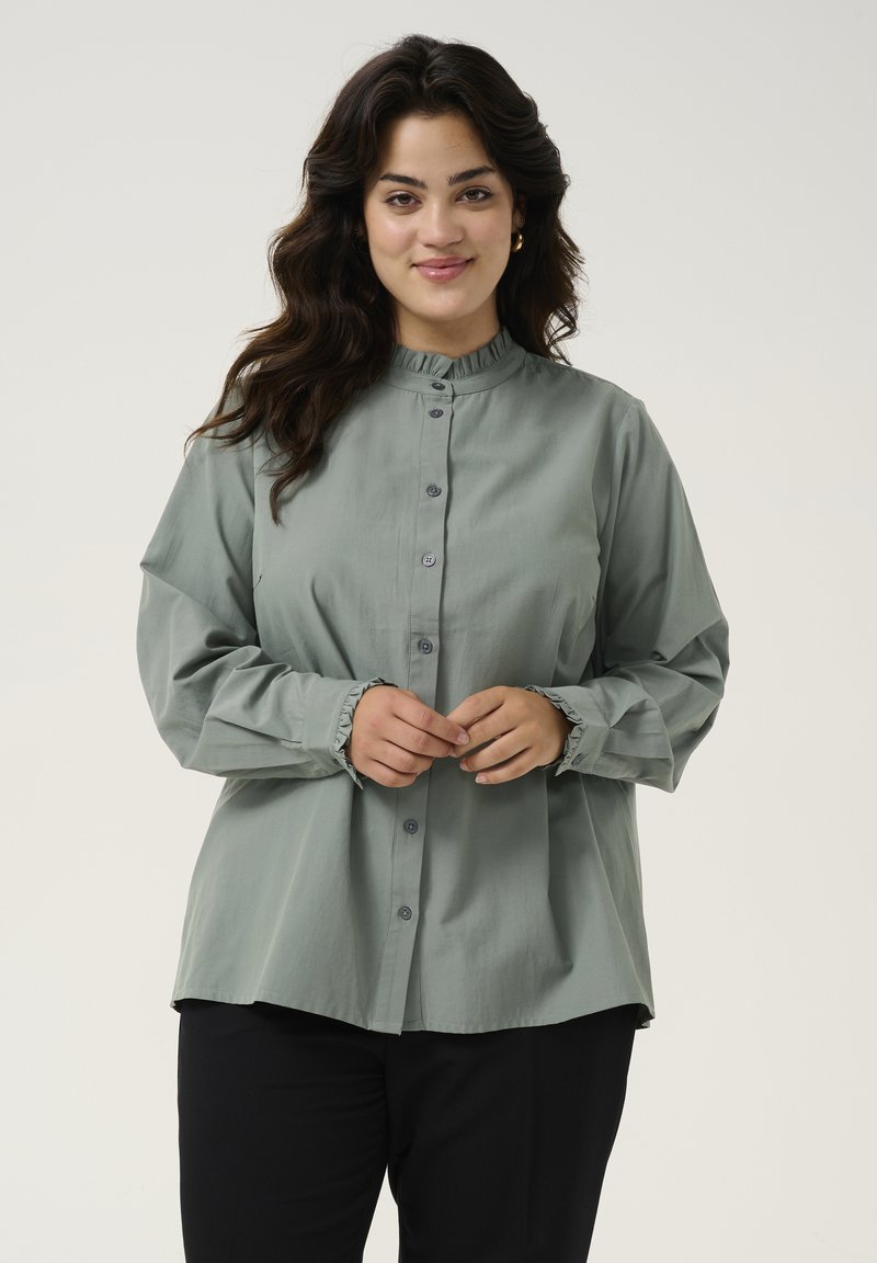 Long-sleeve button-up shirt in light green fabric, featuring a ruffled collar, gray buttons, and gathered cuffs. Worn over black pants.