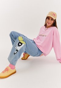 Růžová mikina s vyšívaným designem, světle modré džíny s potiskem karikatury na koleni, žluté boty a béžová kšiltovka.