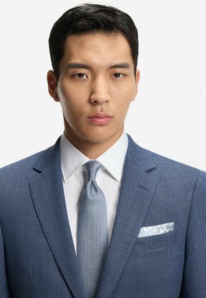 Costume bleu clair texturé avec une chemise blanche, une cravate à motifs et un foulard en soie à motif cachemire. Accent sur la coupe ajustée et les revers structurés.