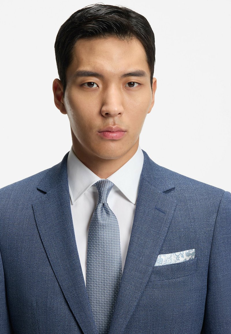 Abito azzurro chiaro con trama, camicia bianca, cravatta a motivo e fazzoletto da taschino paisley. Focus sulla vestibilità sartoriale e sui revers strutturati.