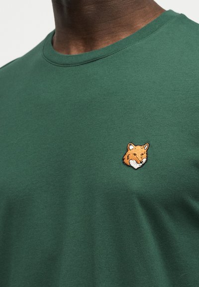 Σκούρο πράσινο μπλουζάκι με μικρό κεντημένο λογότυπο κεφαλής αλεπούς στο αριστερό στήθος, φορεμένο από άτομο με σκουρόχρωμη επιδερμίδα.