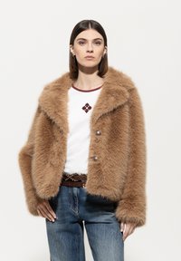 Braune Kunstpelzjacke mit breiten Revers, zwei Frontknöpfen und cropped Schnitt. Getragen über einem weißen Hemd mit burgunderfarbigem Ausschnitt und Akzent.