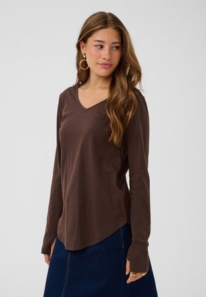 Young woman with long wavy hair wearing large hoop earrings, a brown long-sleeve V-neck shirt, and a dark blue skirt standing against a plain background.