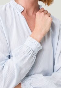 Helles blaues Hemd mit vertikalen weißen Streifen, das einen Rüschensaum am Kragen und an den Manschetten mit gerafftem Detail aufweist. Weicher Stoff und lockere Passform.
