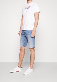 Camiseta blanca de manga corta con rayas horizontales de colores, combinada con pantalones cortos de mezclilla azul claro y zapatillas blancas. Un atuendo simple y casual.