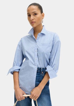 Mujer con camisa de rayas azules abotonada y mangas remangadas, jeans azules, pendiente de aro dorado, sosteniendo el asa de una bolsa blanca.